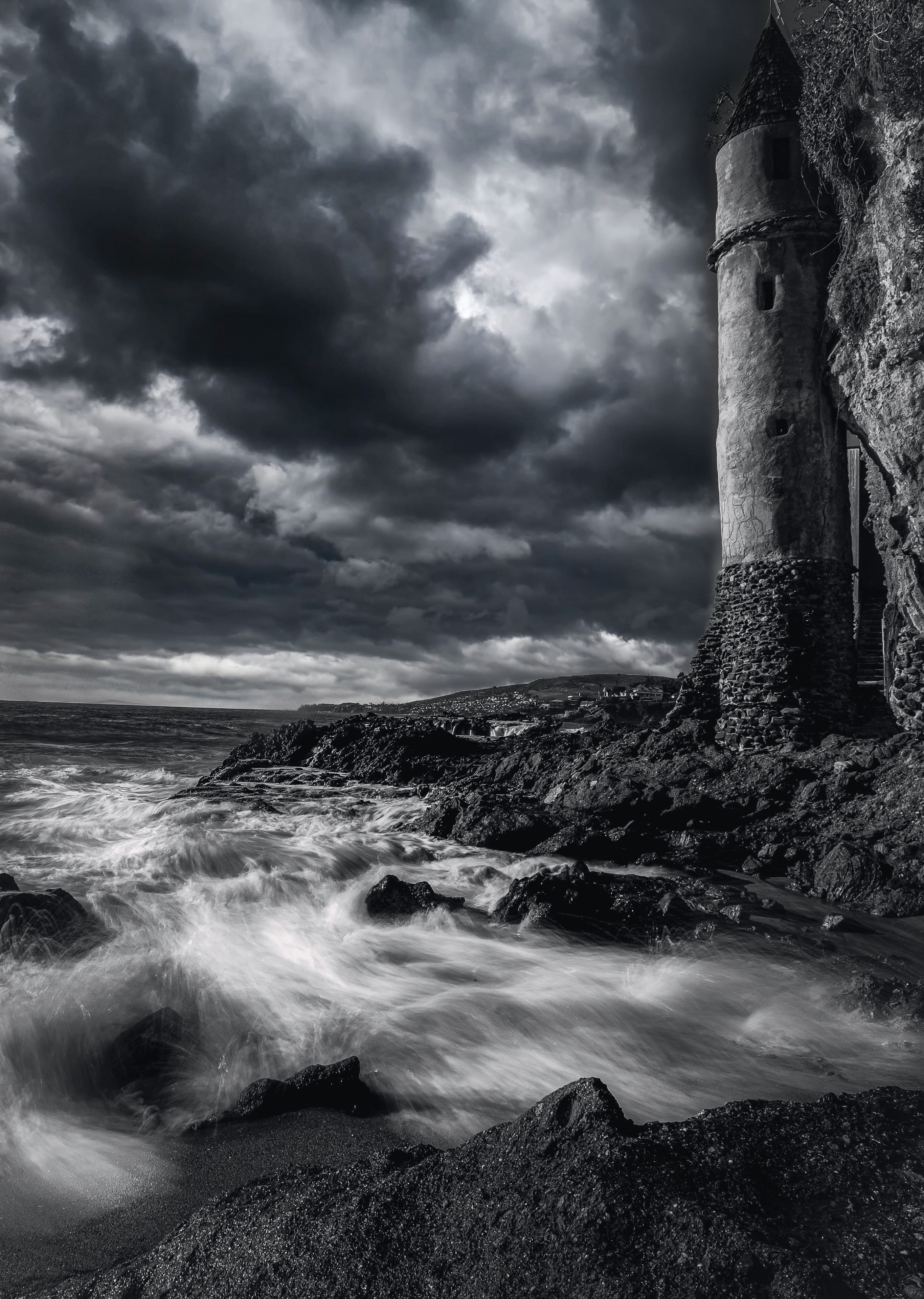 Ruined coastal tower with turbulent waves crashing on rocks under dramatic stormy sky, black and white