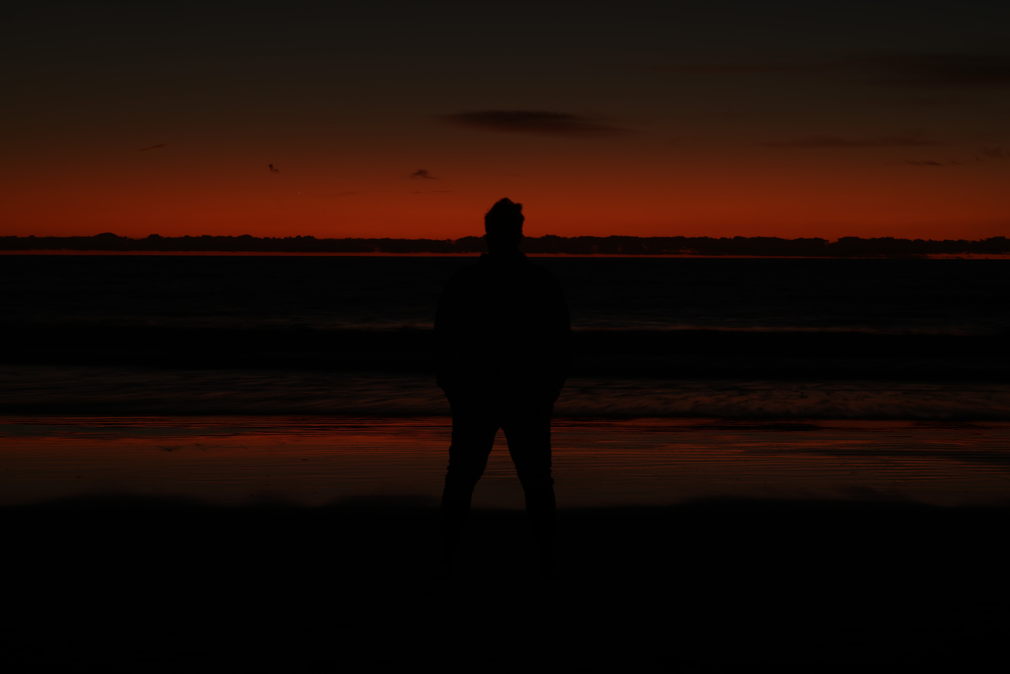 Lone figure silhouetted against deep crimson afterglow reflected on wet tidal flats at dusk