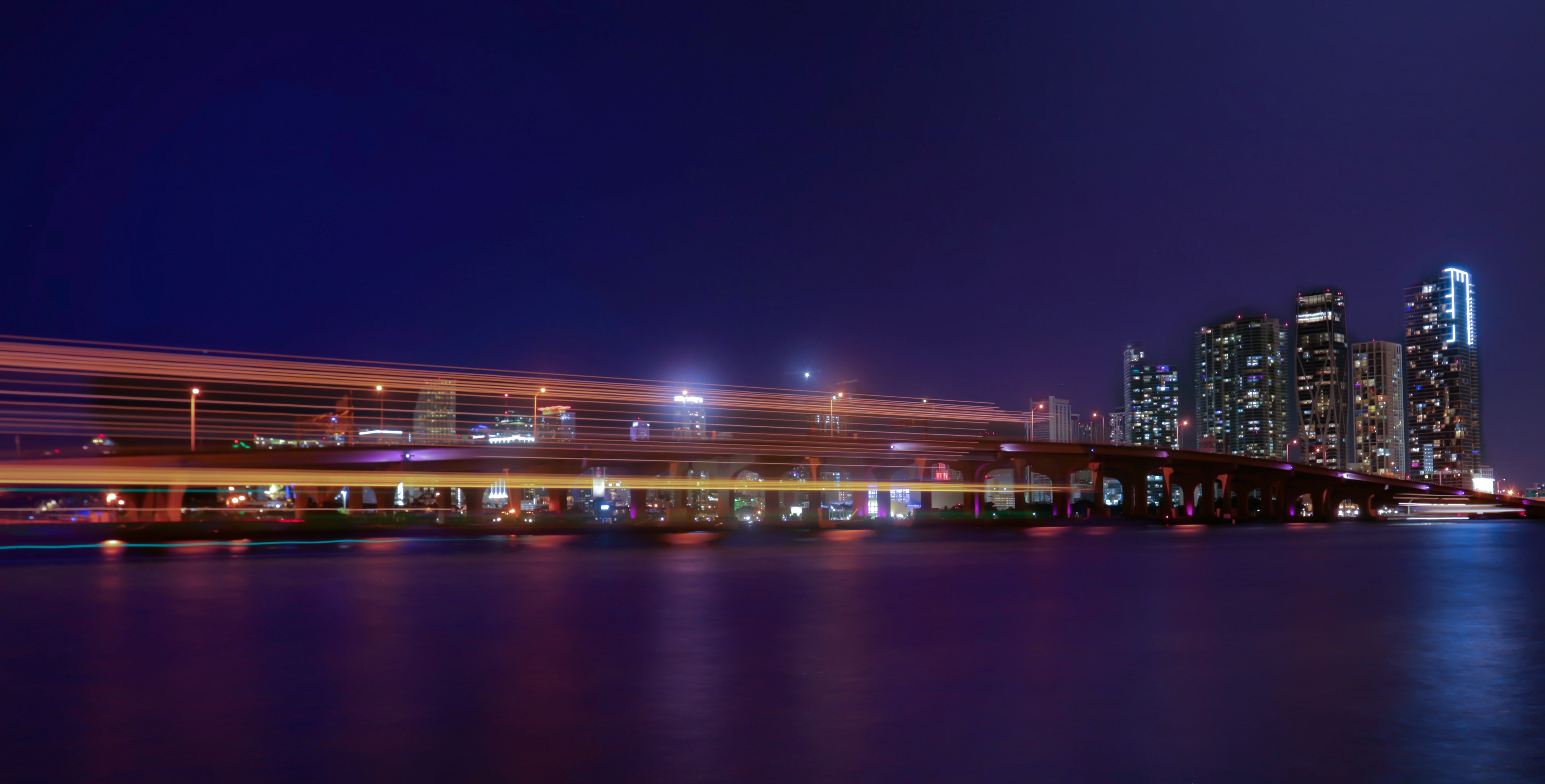 Golden-lit wooden dock extending toward a glittering city skyline reflected in calm dark water at night