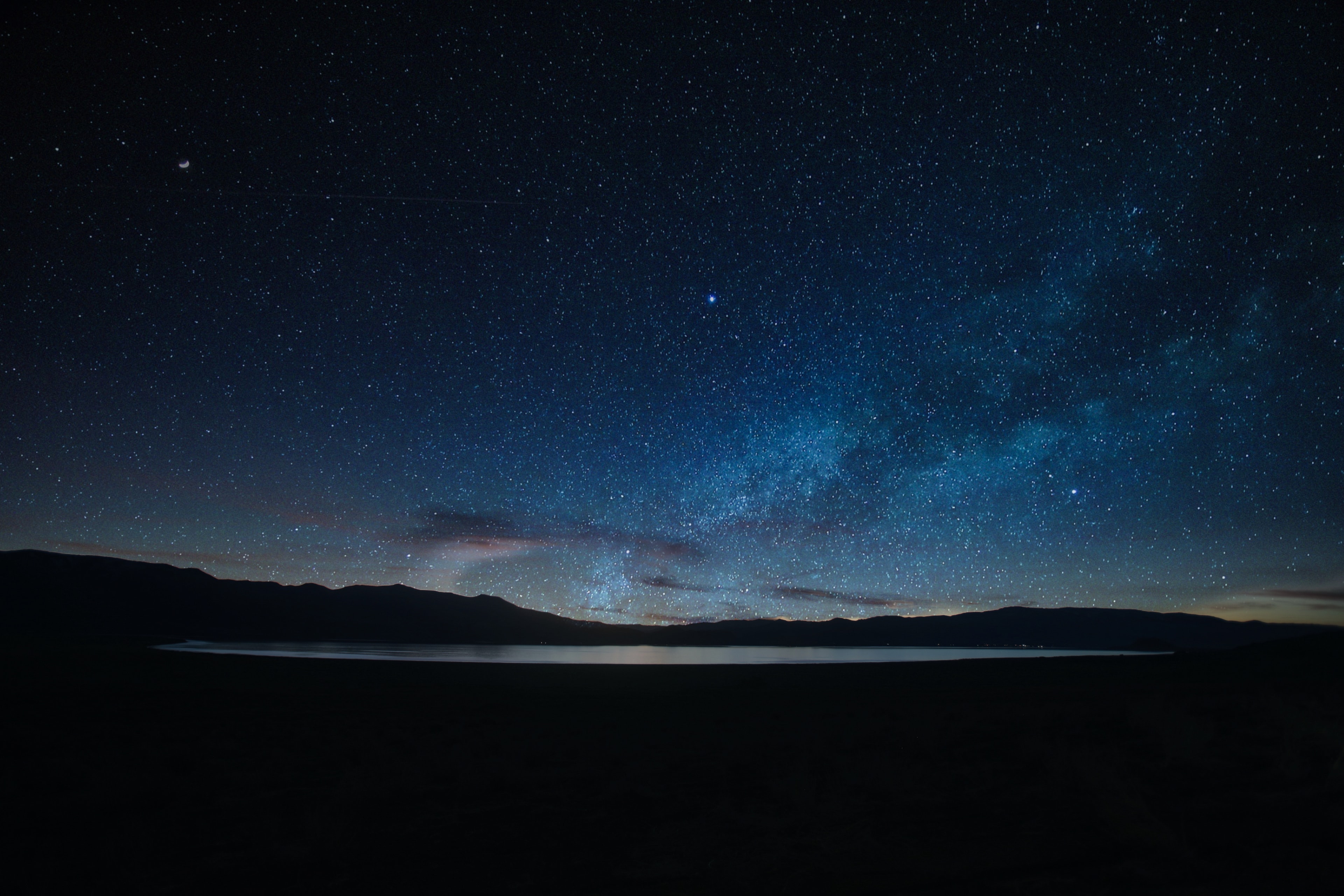Milky Way arching over a dark mountain lake with silhouetted ridgeline and crescent moon against a deep blue starry sky