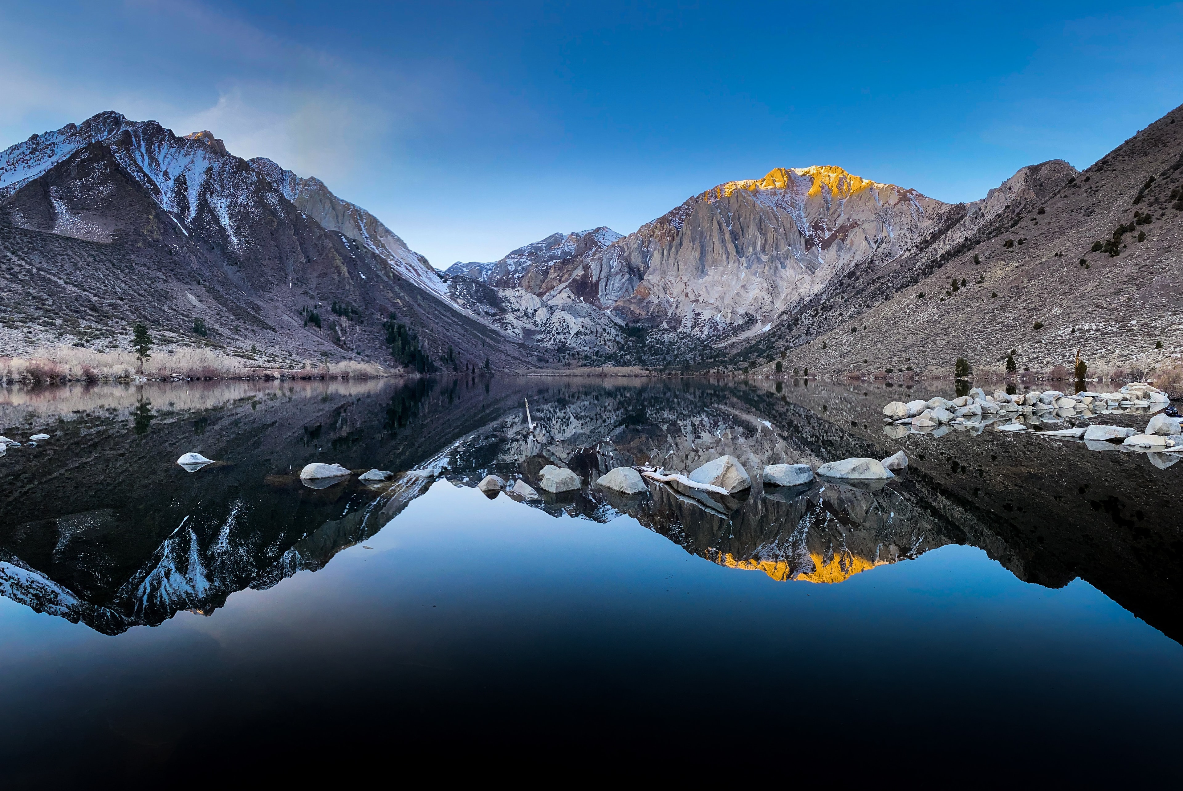 Snow-capped mountain peaks perfectly mirrored in a glassy alpine lake at golden hour with boulders in foreground