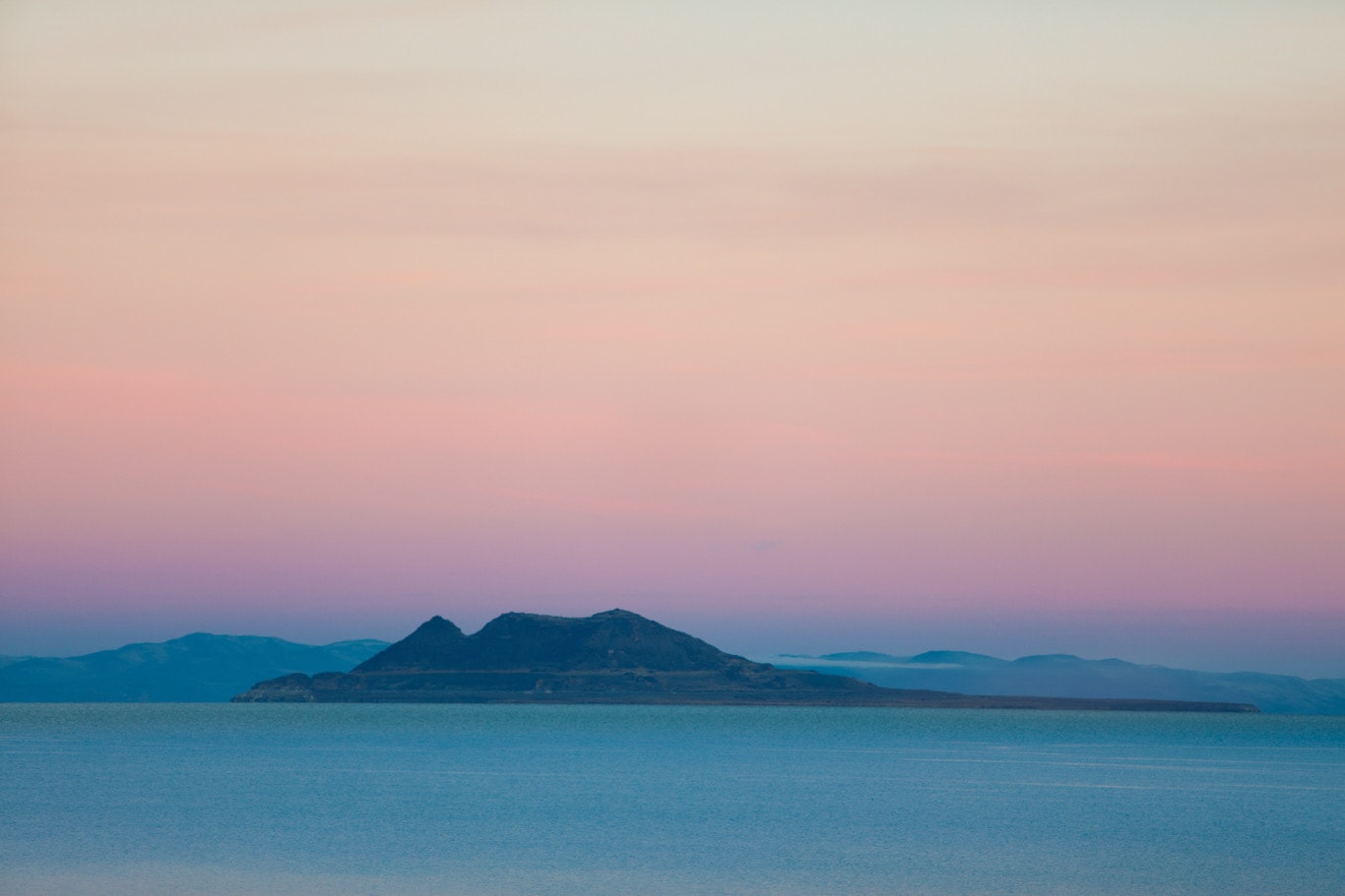 Lone mountain island silhouetted against a soft pink and lavender gradient sky at dusk over glassy calm water